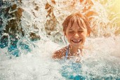 Little girl in waterpark pool being splashed by waterfall. Sunny summer day.
